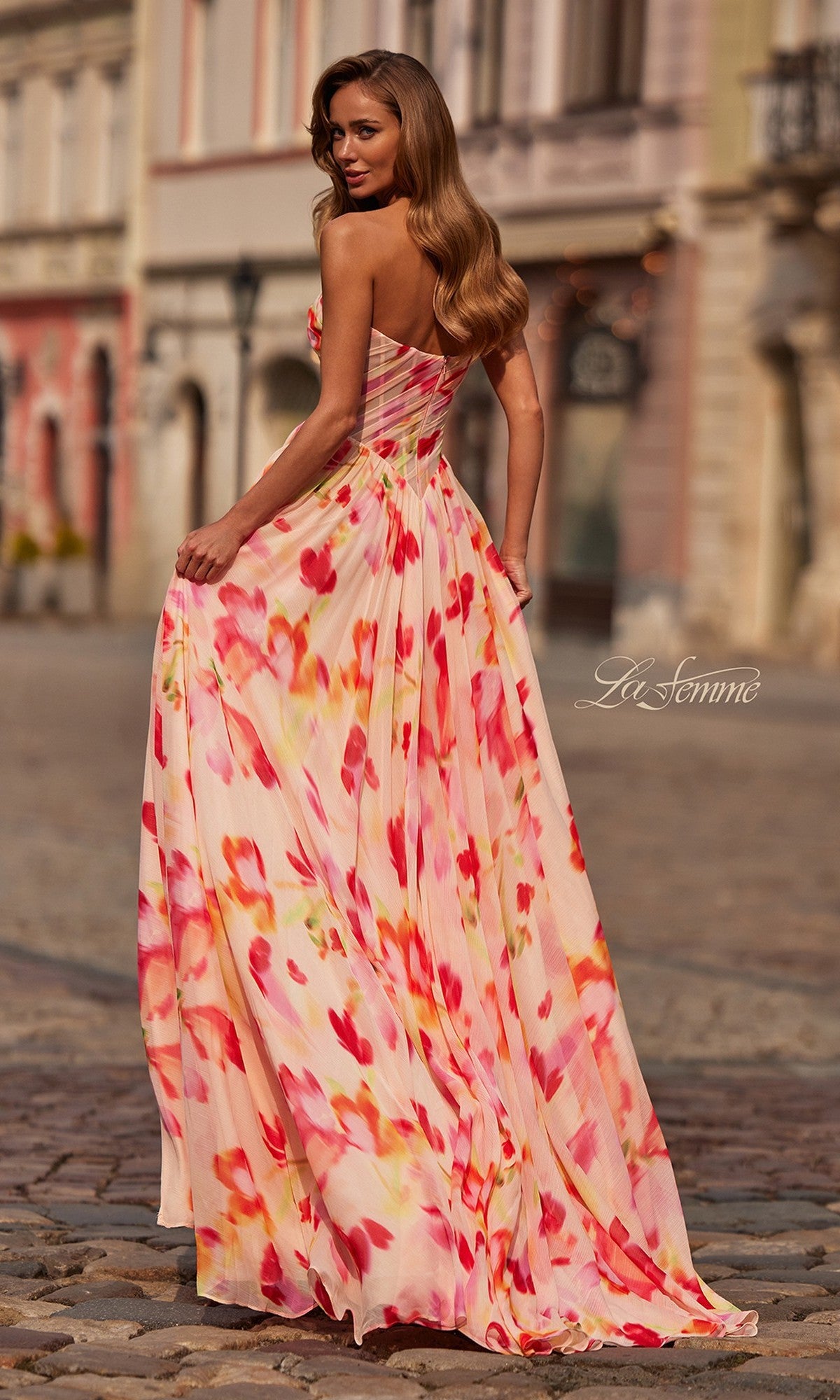 A woman stands on a cobblestone street wearing the La Femme 33404 long prom dress with a strapless pink and orange floral chiffon design, looking back over her shoulder.